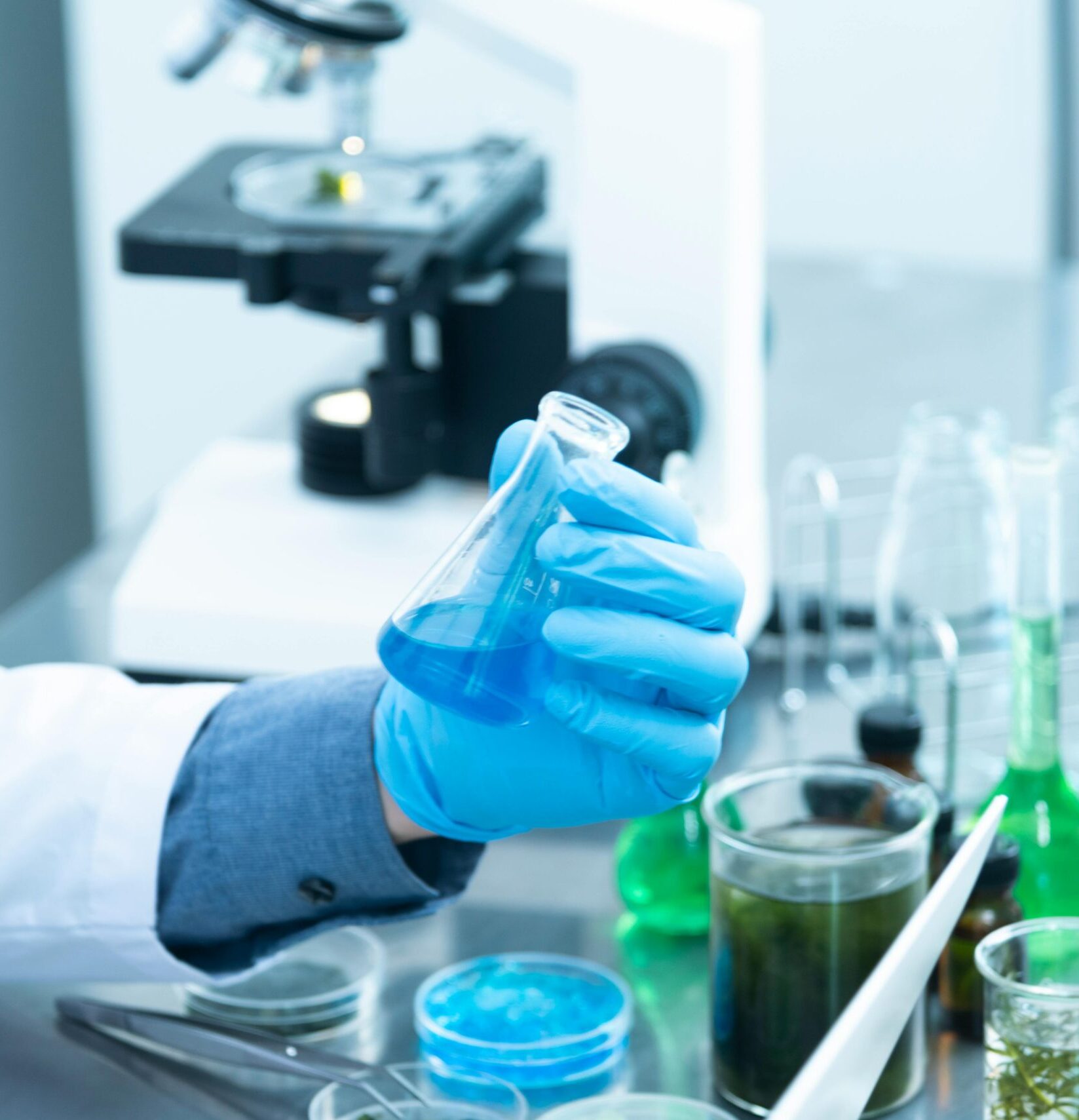 Scientist in gloves analyzing blue liquid in a laboratory setting with microscope and glassware.
